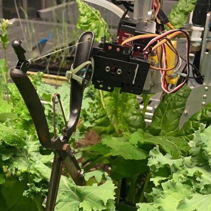 A robotic servo on a gantry, zip-tied to a pair of pruning shears above a green garden plot.