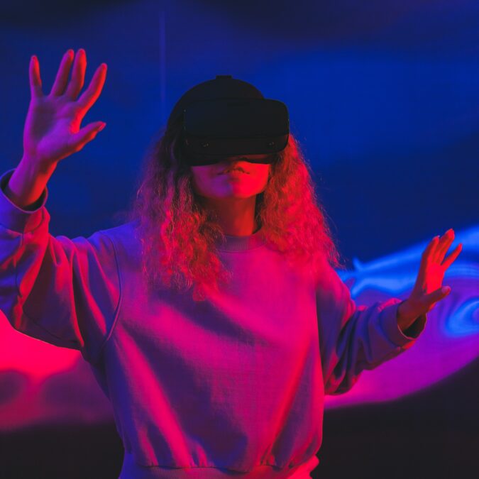 A woman with curly hair in blue and pink lighting wears a VR headset with her arms slightly outstretched.
